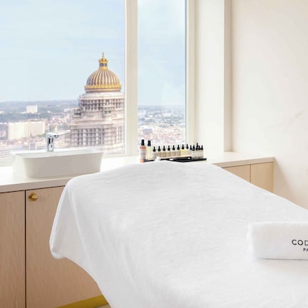 A hotel room with a bed, a window with a view of a cityscape and a large building, and toiletries on the bedside table.