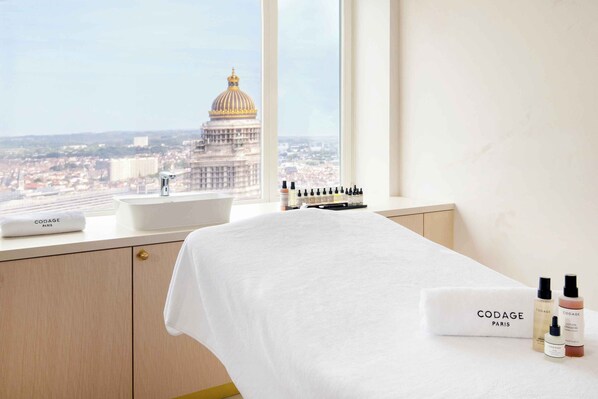 A hotel room with a bed, a window with a view of a cityscape and a large building, and toiletries on the bedside table.