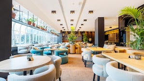 Un restaurant modern amb taules i cadires rodones, una paret de vidre amb plantes en maceta i una zona de seients acollidora amb sofàs i coixins.