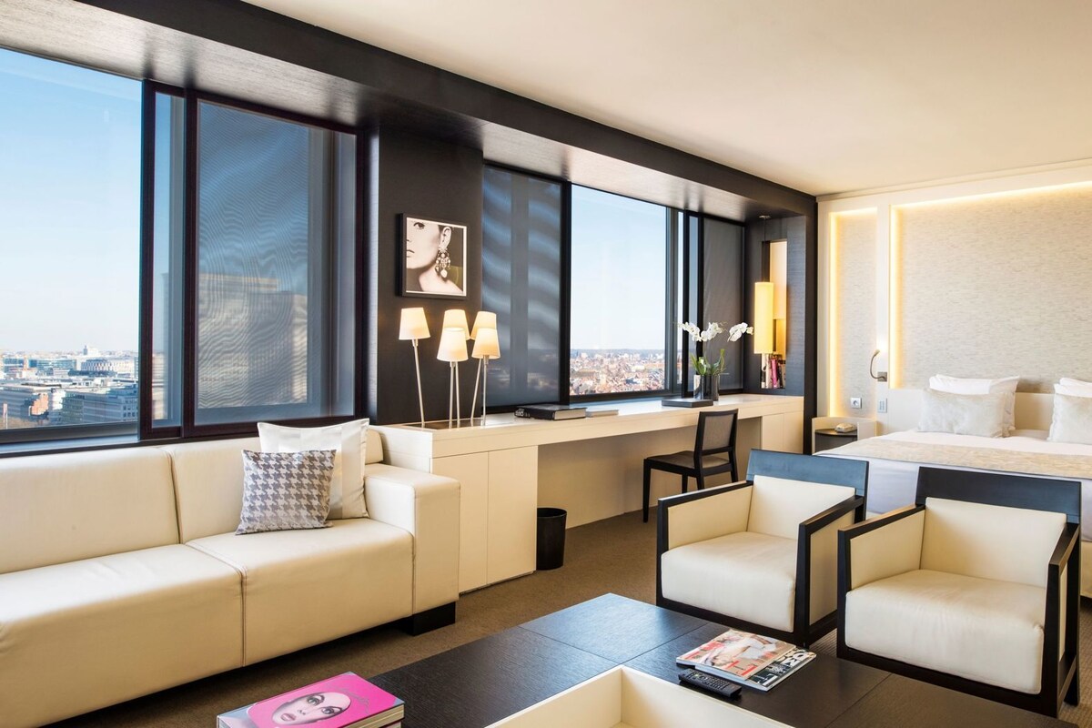 Una habitación de hotel moderna con una cama grande, un sofá, dos sillones, un escritorio y un amplio ventanal con vistas a la ciudad.