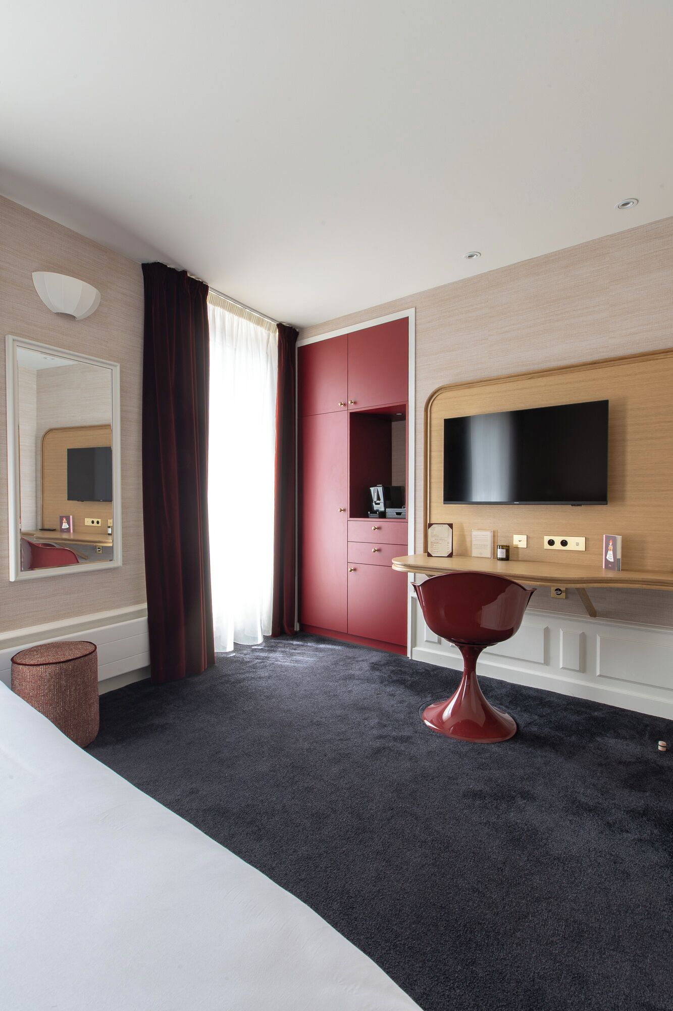 A hotel room with a bed, a desk, a television, a mirror, and a red wardrobe.