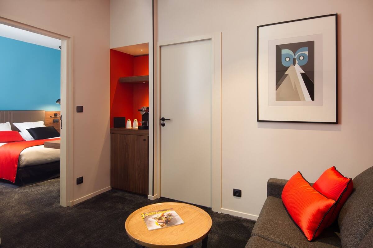Habitación de hotel con cama, una mesita, sofá con cojines naranjas y una obra de arte enmarcada en la pared.