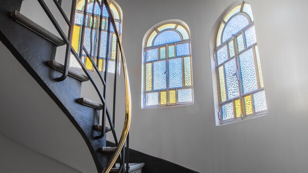 A spiral staircase with a black handrail and patterned steps, leading to a landing with two arched windows.