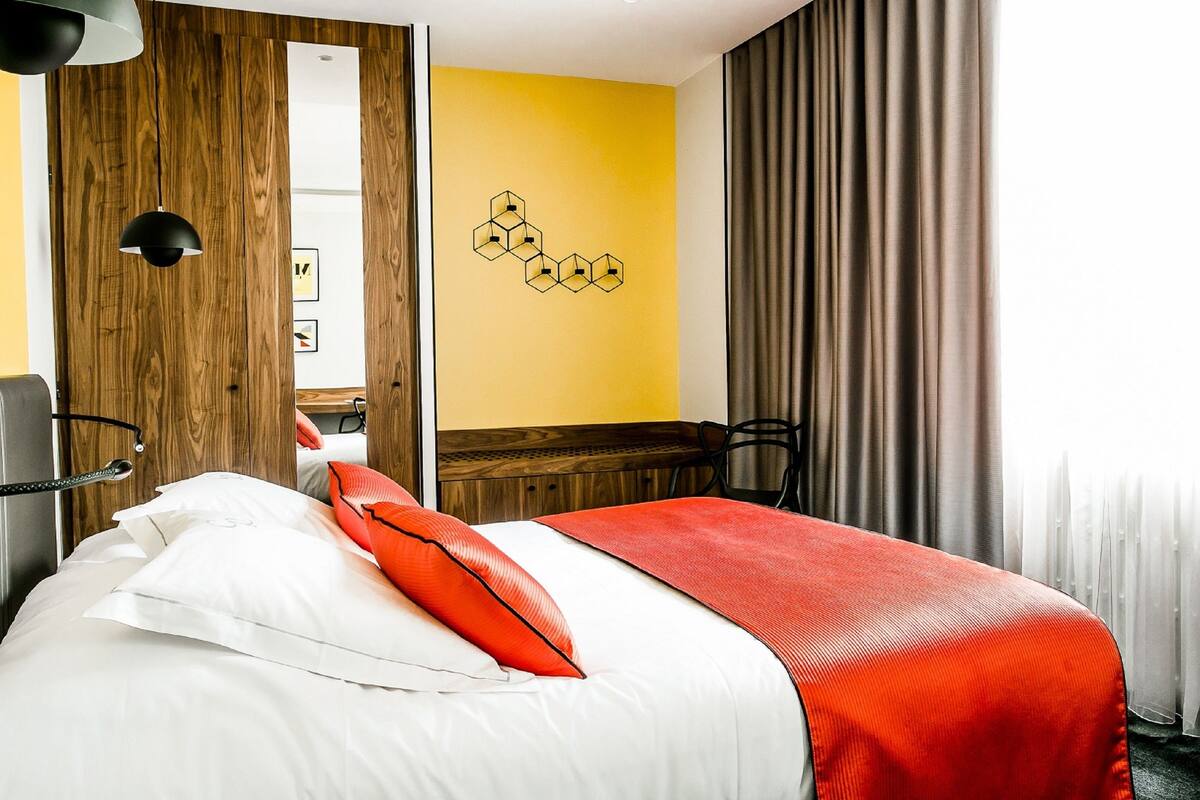 Habitación de hotel con cama, armario de madera, pared amarilla y ventana con cortinas.