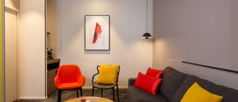 A modern living room with a grey sofa, orange and yellow chairs, a wooden coffee table, and a framed artwork on the wall.