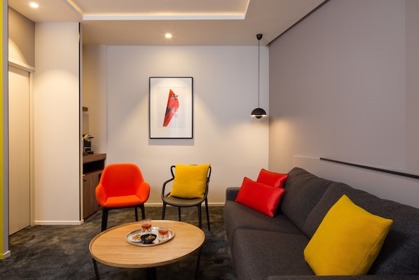 A modern living room with a grey sofa, orange and yellow chairs, a wooden coffee table, and a framed artwork on the wall.