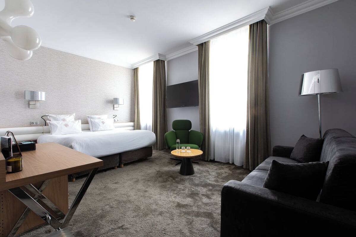 Una habitación de hotel con una cama, un sillón verde, una mesita redonda pequeña, un escritorio con una botella y un televisor montado en la pared.