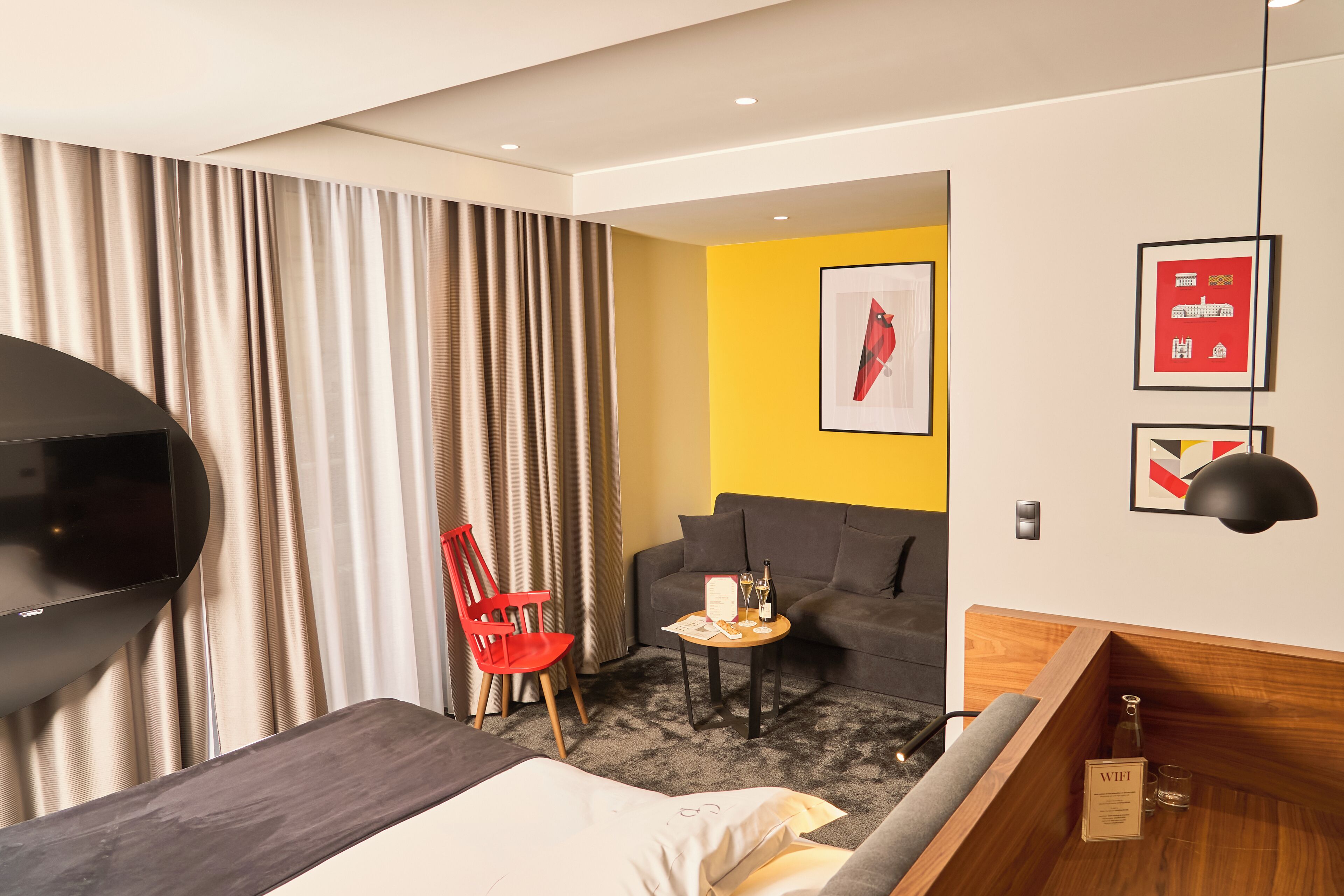 A hotel room with a bed, a TV, a red chair, a small table with a bottle, a sofa, and a yellow accent wall.