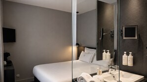 A modern hotel room with a bed, a glass partition, a bedside table with a coffee maker, a mirror with toiletries, and a sink.