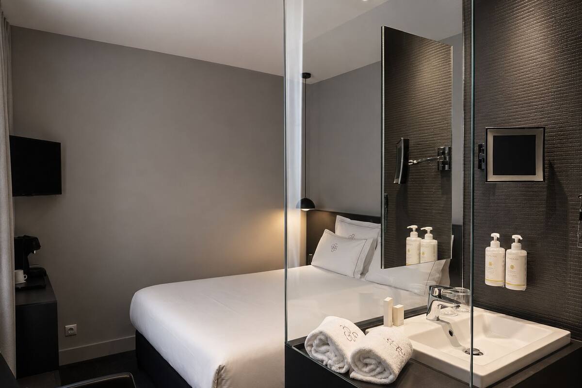 Una habitación de hotel moderna con una cama, un separador de vidrio, una mesita de noche con cafetera, un espejo con artículos de aseo y un lavamanos.