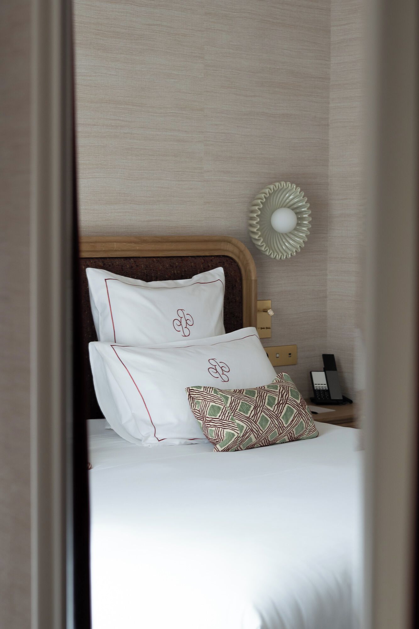 Una cama bien hecha con ropa de cama blanca y almohadas estampadas, una mesita de noche con teléfono y lámpara, y una lámpara de pared.