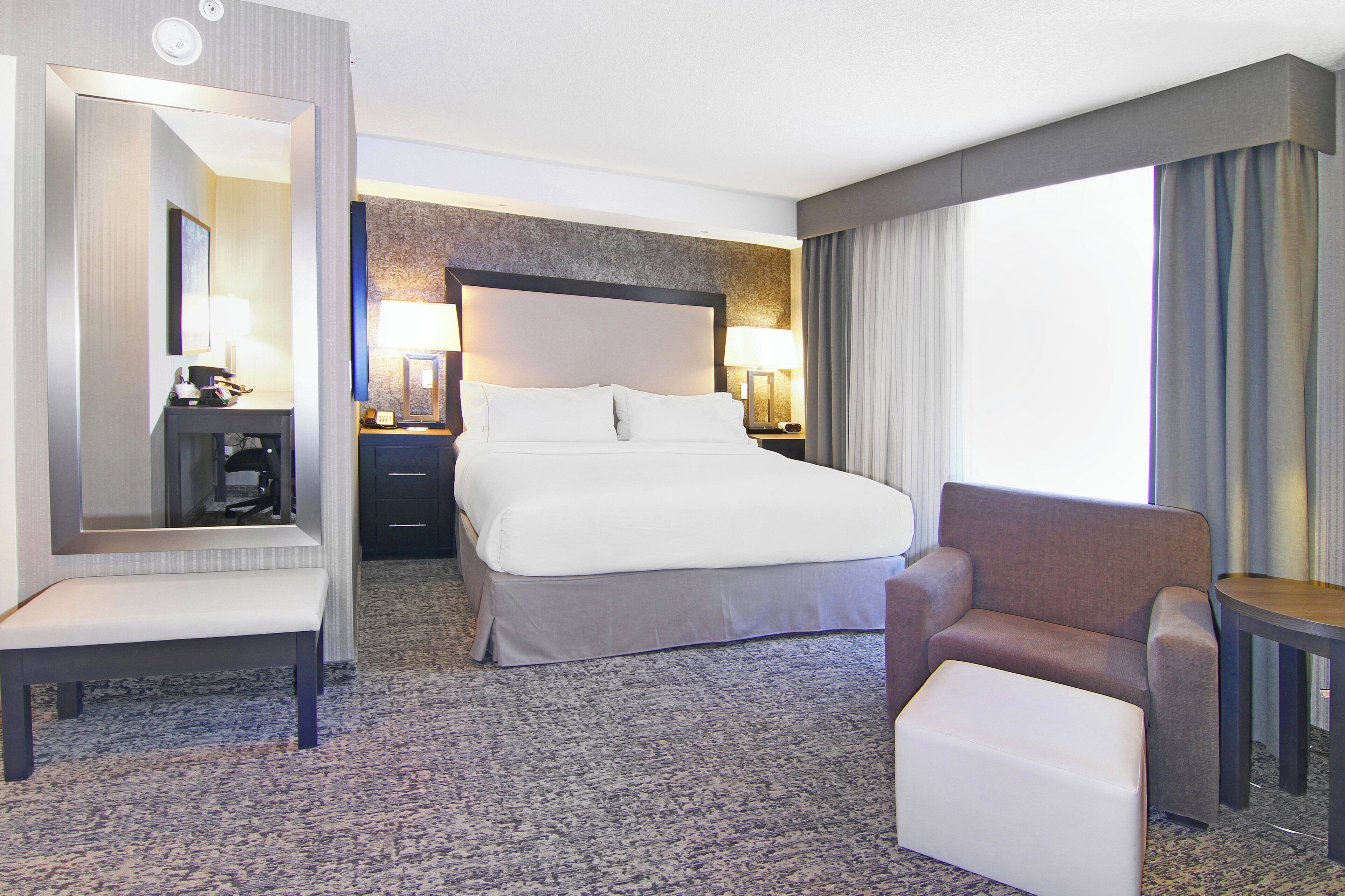 Habitación de hotel con una cama grande, una silla, una mesita, un espejo y una mesita de noche con lámparas.