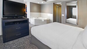 A modern hotel room with a flat-screen TV, a large bed, a bathroom with a bathtub, and a mirror reflecting the room. - Holiday Inn Express Hotel & Suites Calgary by IHG (Calgary)