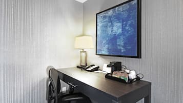 A hotel room with a desk, a lamp, a coffee maker, and a monitor.