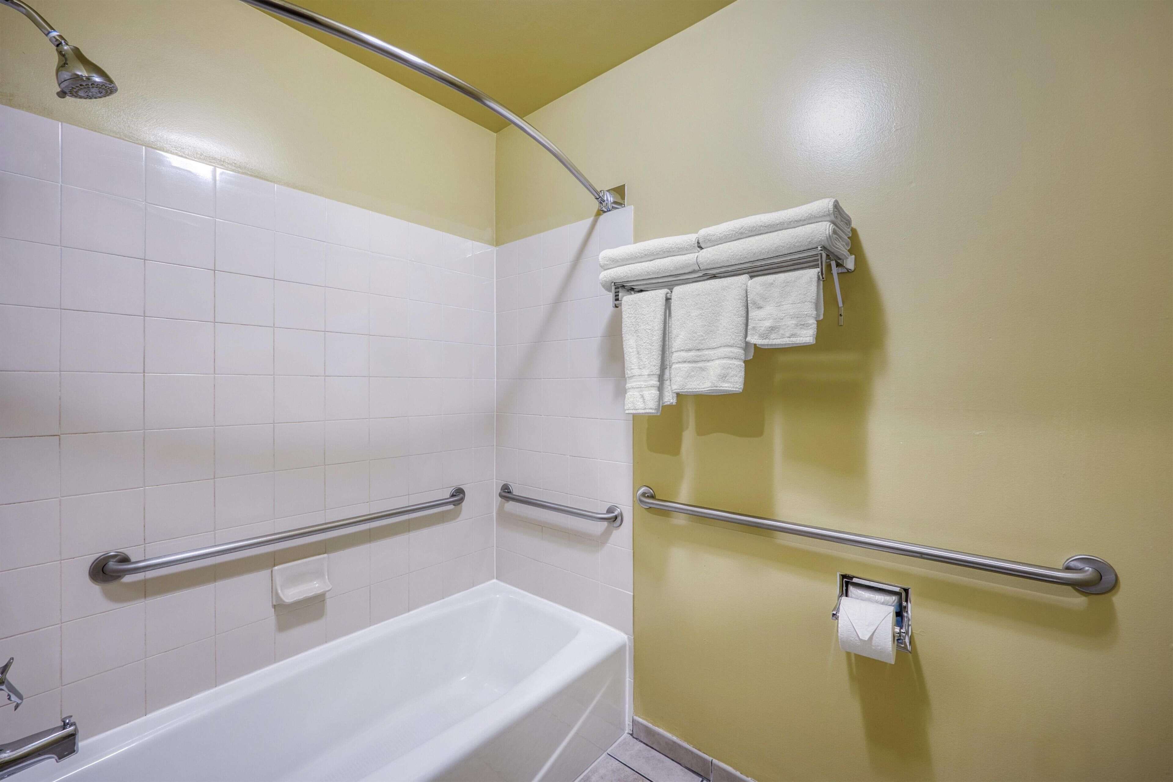 Un baño con una bañera blanca, barras de apoyo, un perchero con toallas y un dispensador de papel higiénico.