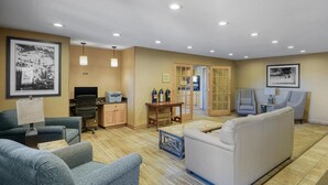 Ein Hotelzimmer mit Wohnbereich, ausgestattet mit einer Couch, Sesseln, einem Couchtisch, einem Fernseher und einem Schreibtisch mit einem Computer. An den Wänden hängen gerahmte Bilder und eine Tür führt in einen weiteren Raum.