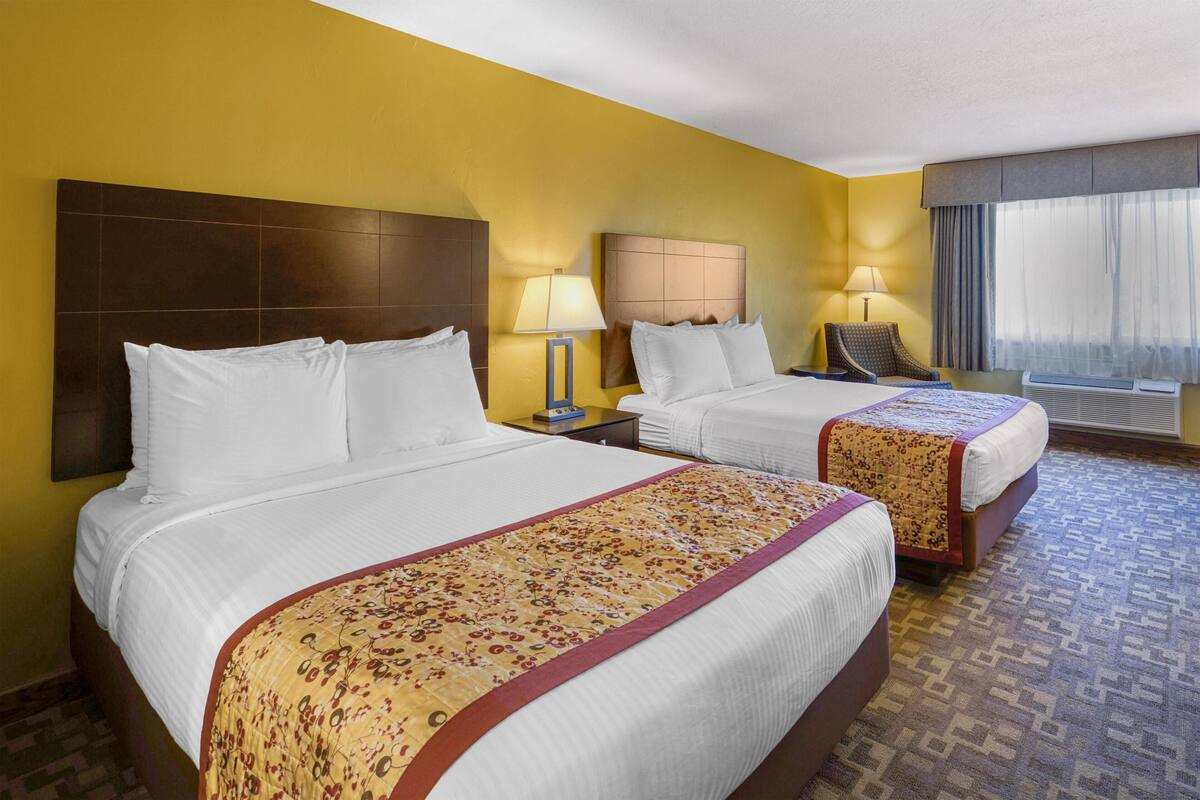 A hotel room with two beds, a nightstand, a lamp, a chair, and a window with curtains.