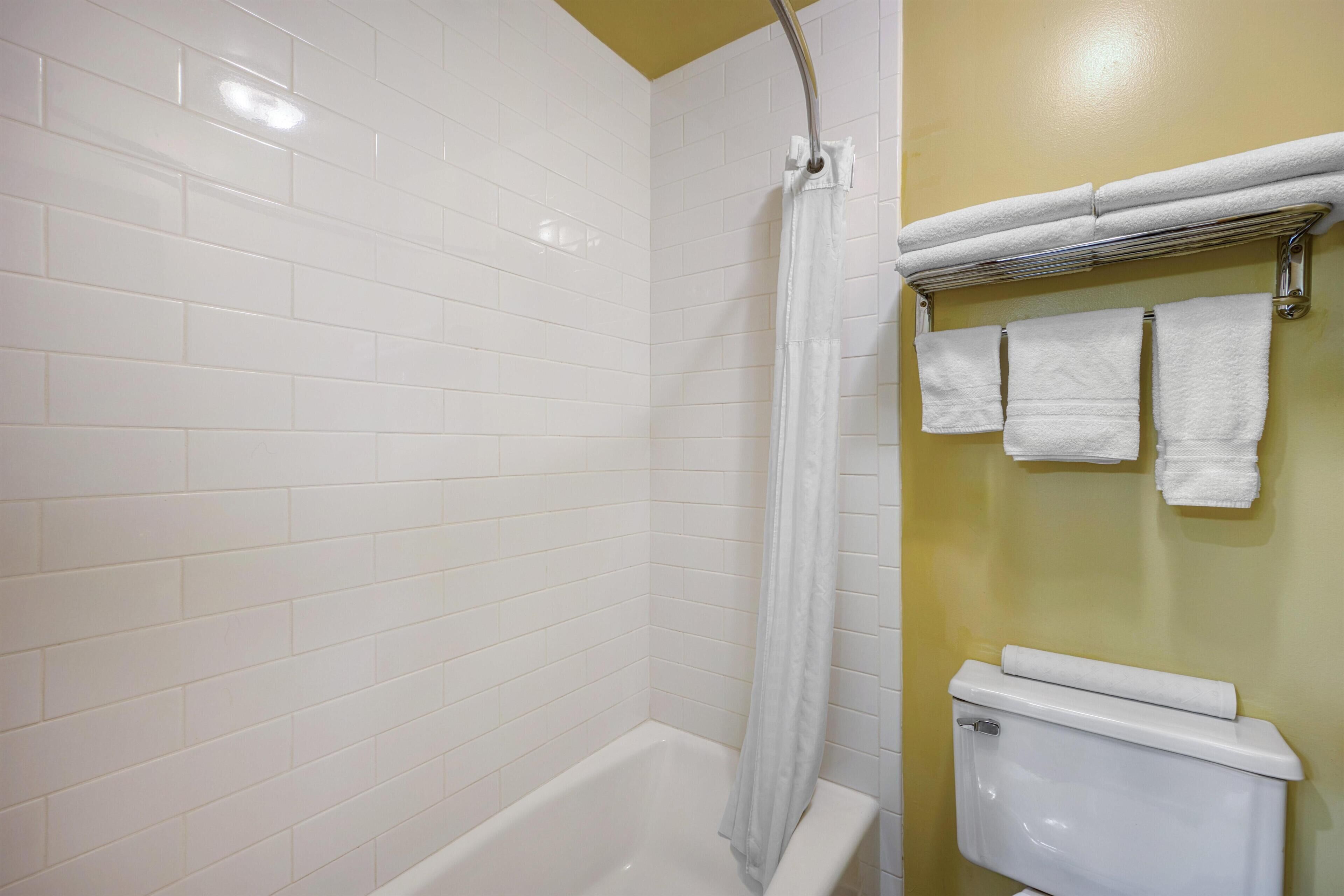 Un baño con paredes cubiertas de azulejos blancos, una bañera blanca, una cortina de ducha y un inodoro con un toallero encima.