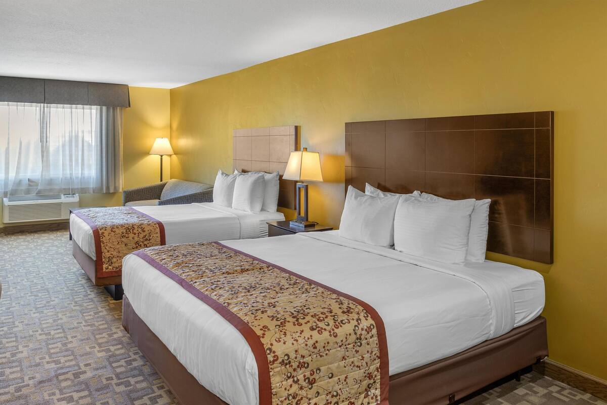 A hotel room with two beds, a large window with curtains, a standing lamp, and a headboard.