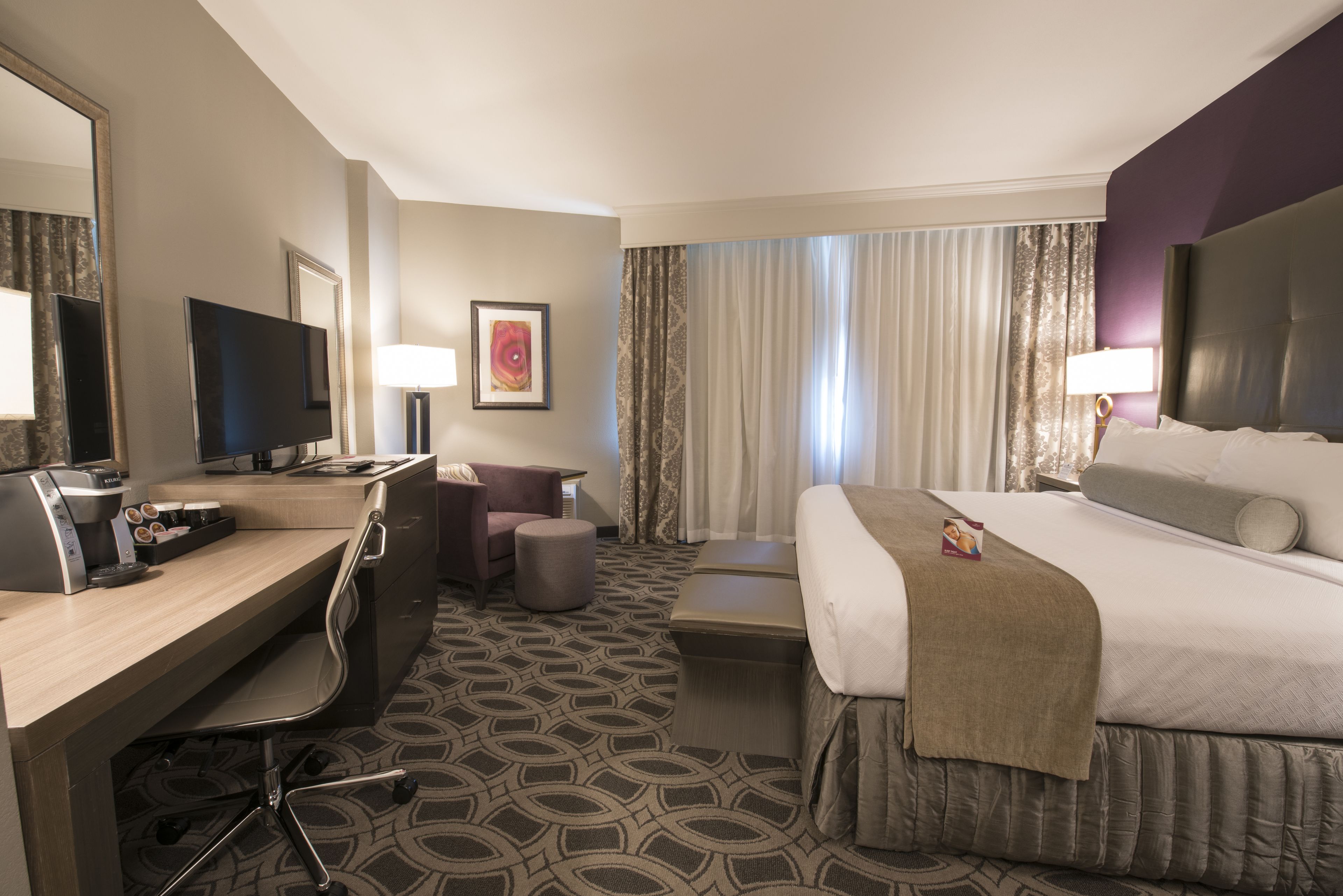A hotel room with a desk, chair, television, bed, and a large window with curtains.