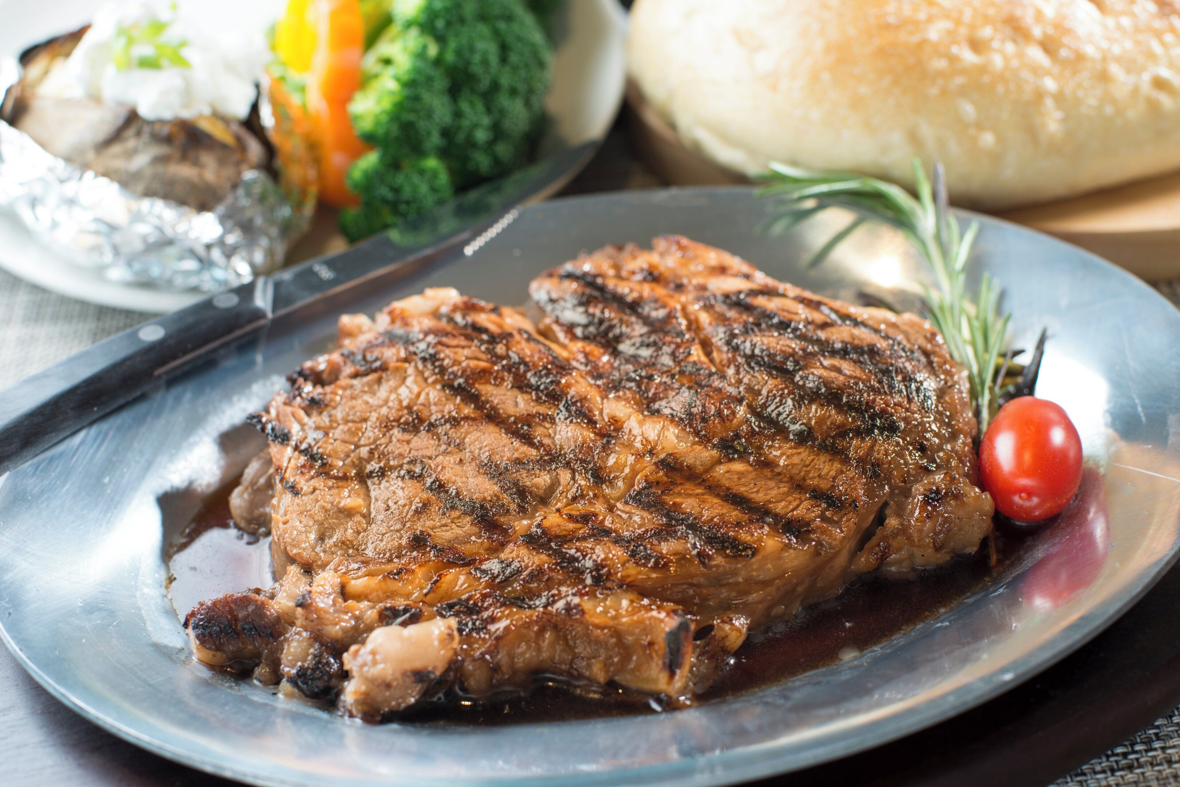A steak with grill marks on a plate, accompanied by a side of vegetables and a roll.