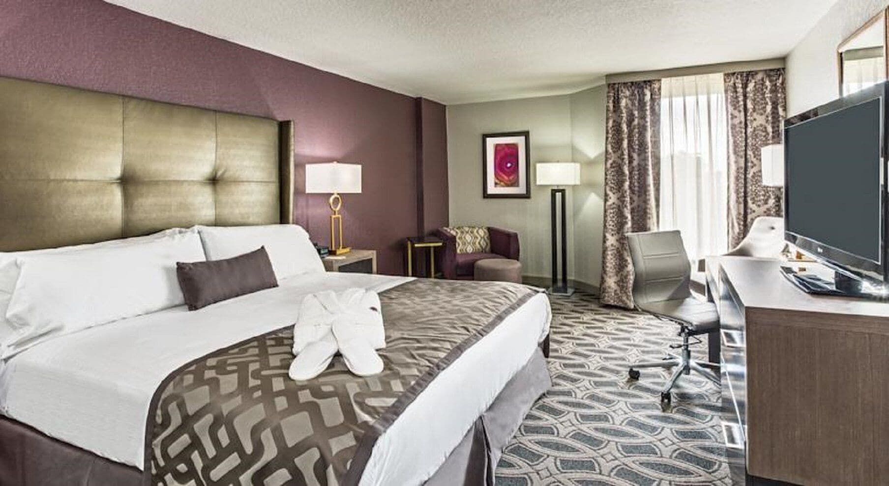 Ein Hotelzimmer mit einem großen Bett, einem Schreibtisch mit Flachbildfernseher, einem Sessel und einem kleinen Tisch mit einer Lampe.
