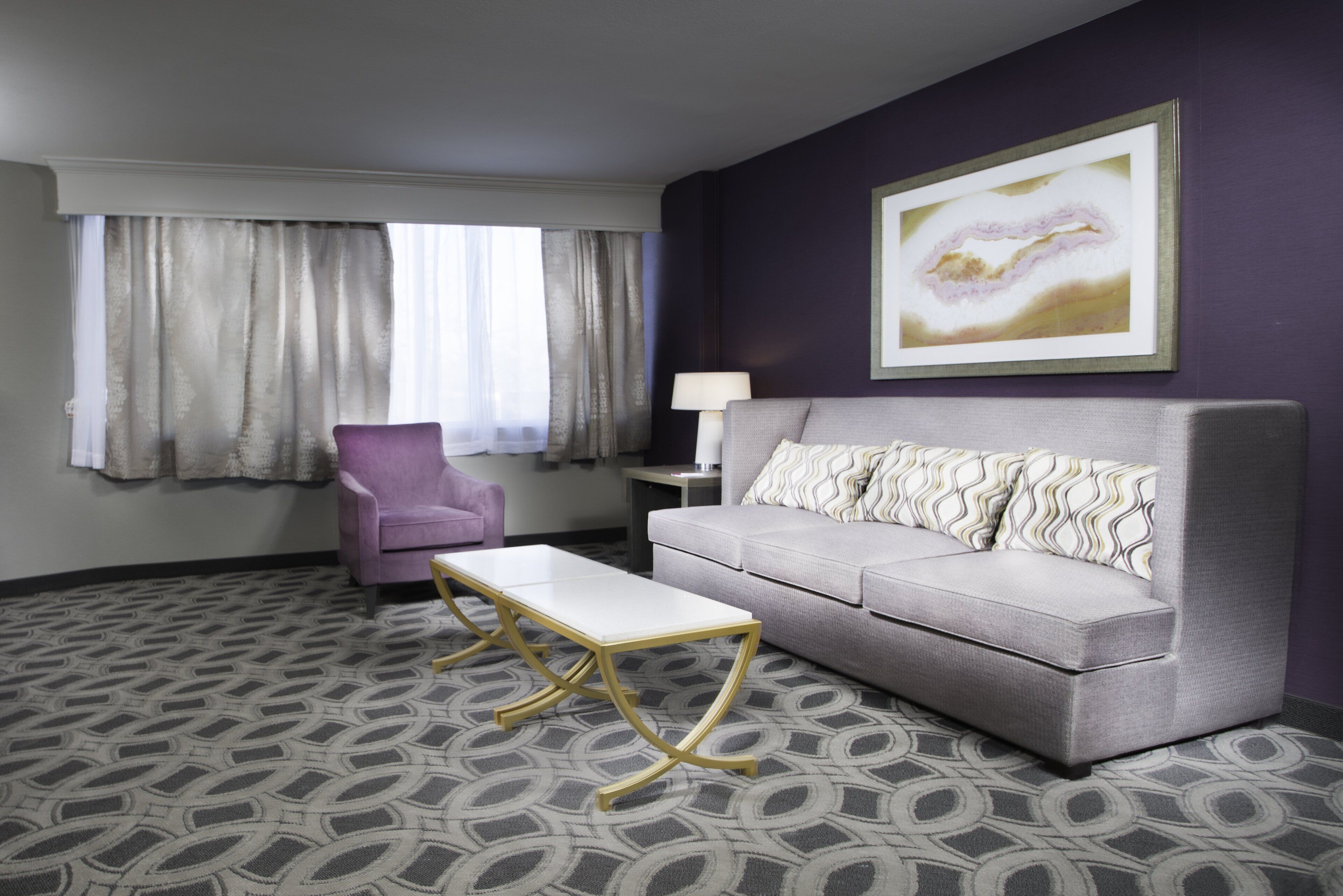 A hotel room with a grey sofa, a white coffee table, a purple armchair, a side table with a lamp, and a large abstract painting on the wall.