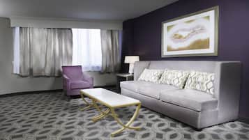 A hotel room with a grey sofa, a white coffee table, a purple armchair, a side table with a lamp, and a large abstract painting on the wall.