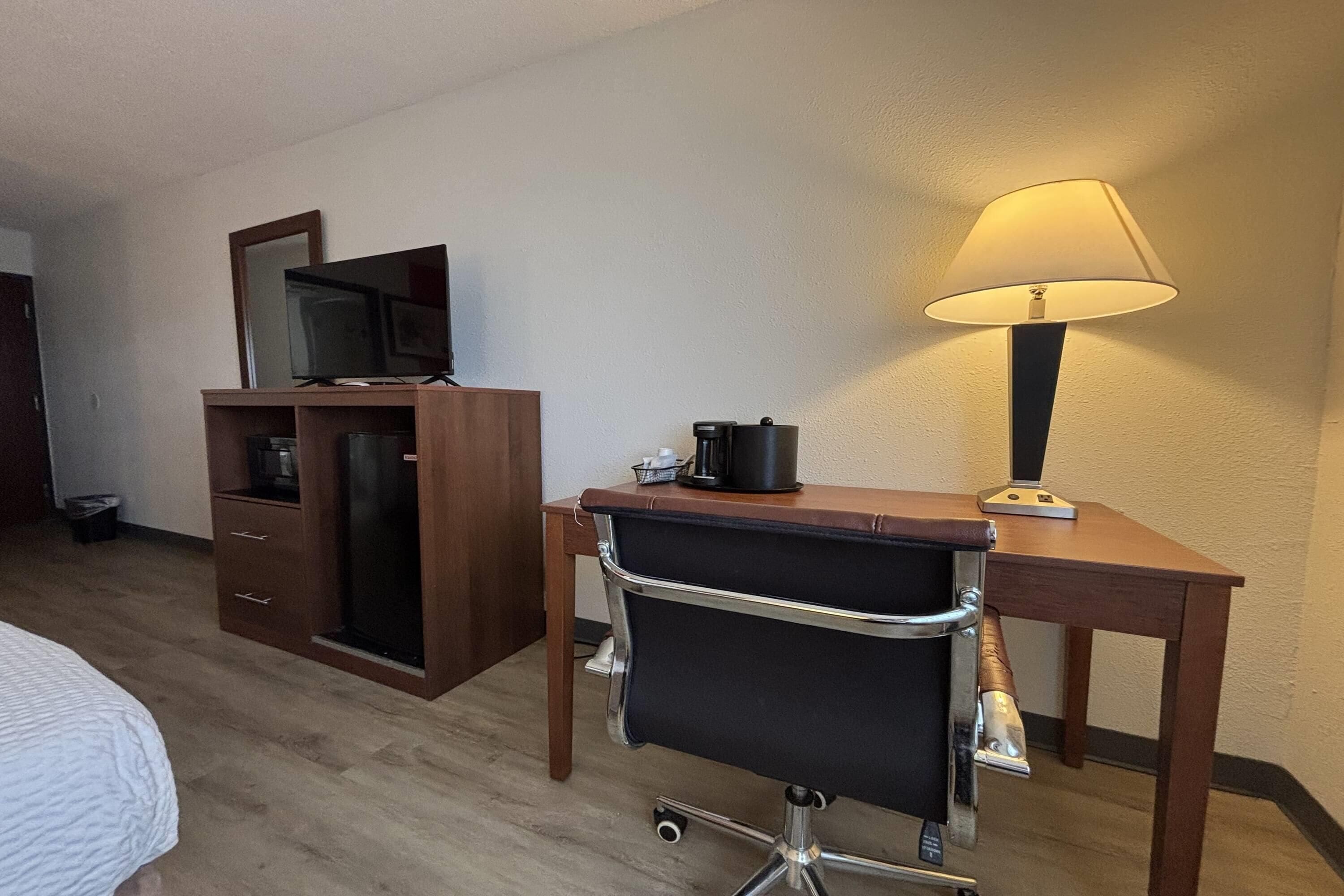 A hotel room with a bed, a desk with a chair, a lamp, a television on a cabinet, and a coffee maker.