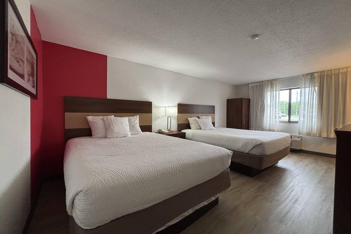 A hotel room with two beds, a red accent wall, a window with curtains, and a wooden floor.