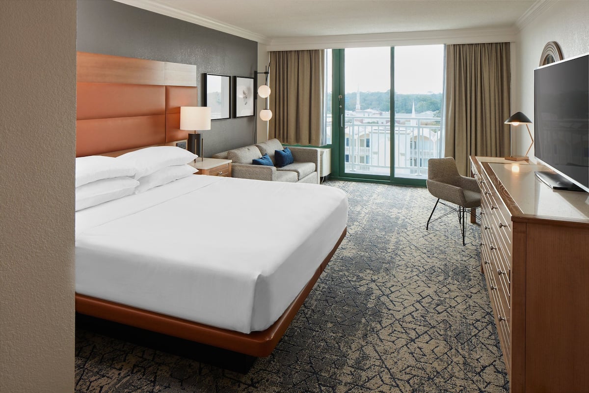 A hotel room with a large bed, a desk with a TV, a sofa, and a view of a cityscape through the window.