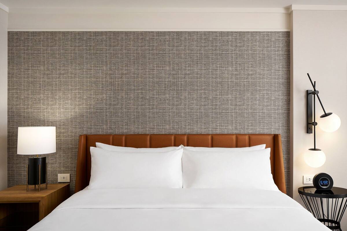 A hotel room with a large bed, a nightstand with a lamp, and a wall clock.