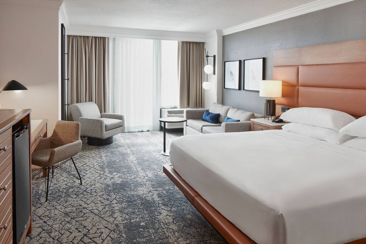 A hotel room with a large bed, a sofa, a desk, and a chair. There is a window with curtains, a lamp, and a picture on the wall.