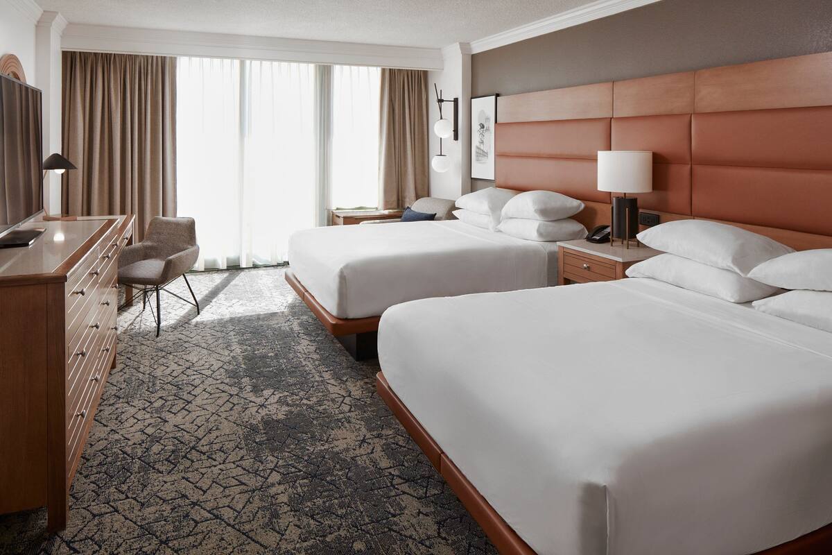 A hotel room with two beds, a desk, a chair, a lamp, and a large window with curtains.