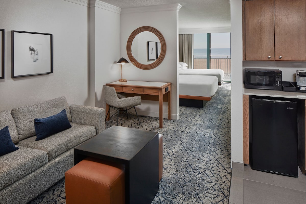 A hotel room with a sofa, a desk, a chair, a bed, a small kitchenette, and a view of the sea.