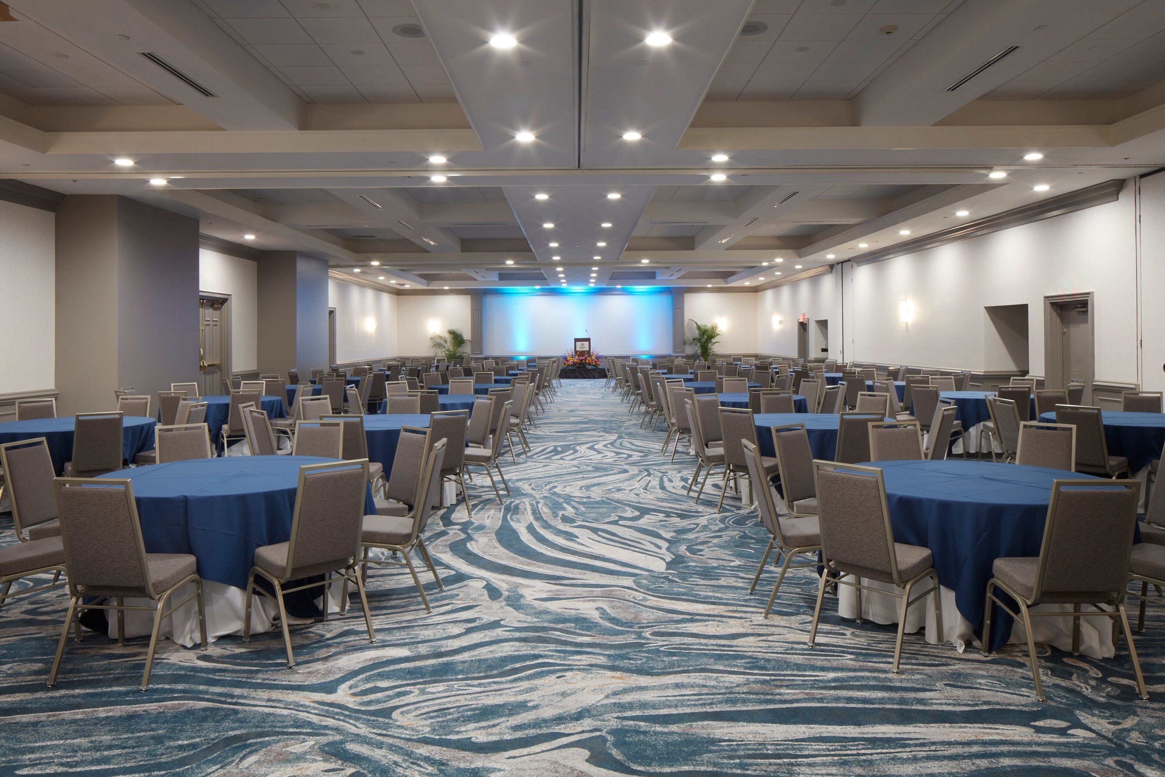 Une grande salle de conférence avec des rangées de tables et de chaises, une scène à l’avant et un tapis bleu orné d’un motif ondulé.