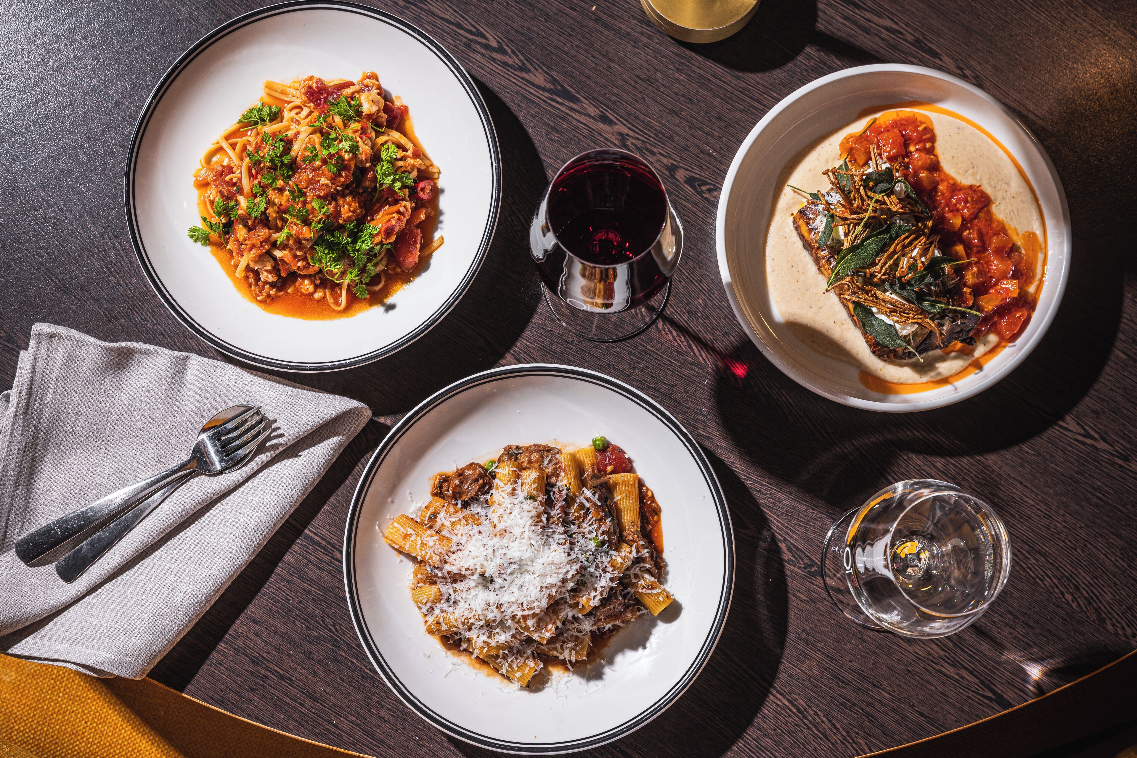 Tres platos de pasta sobre una mesa de madera, con un tenedor y una servilleta a la izquierda, una copa de vino tinto en el centro y una copa de agua a la derecha.