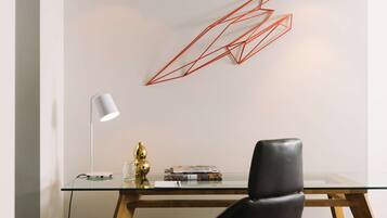 Un espacio de oficina moderno con un escritorio de vidrio, una silla de cuero negro, una lámpara de escritorio blanca y un adorno rojo geométrico en la pared.