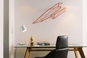 Un espacio de oficina moderno con un escritorio de vidrio, una silla de cuero negro, una lámpara de escritorio blanca y un adorno rojo geométrico en la pared.