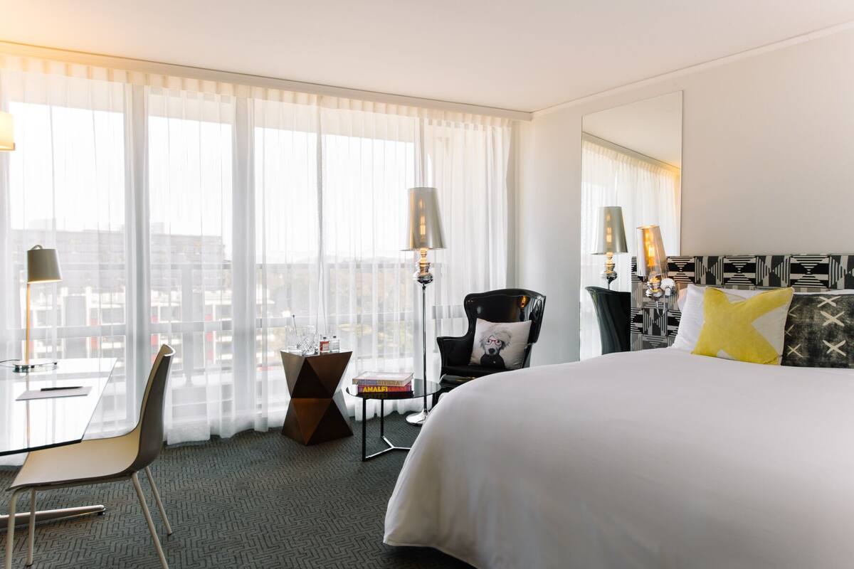 Quarto de hotel com cama grande, escrivaninha, cadeira e vista da cidade através de cortinas leves.