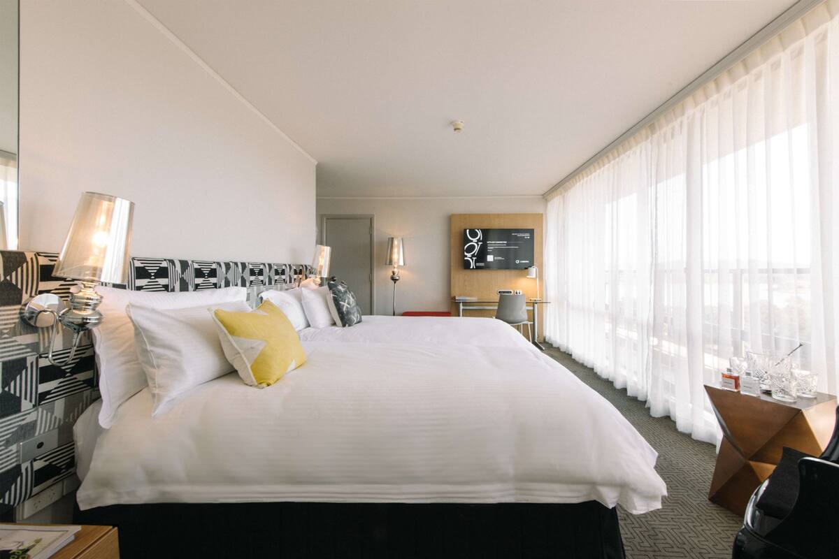 Quarto de hotel com uma cama grande, uma escrivaninha, uma cadeira, uma TV e uma janela com cortinas translúcidas.