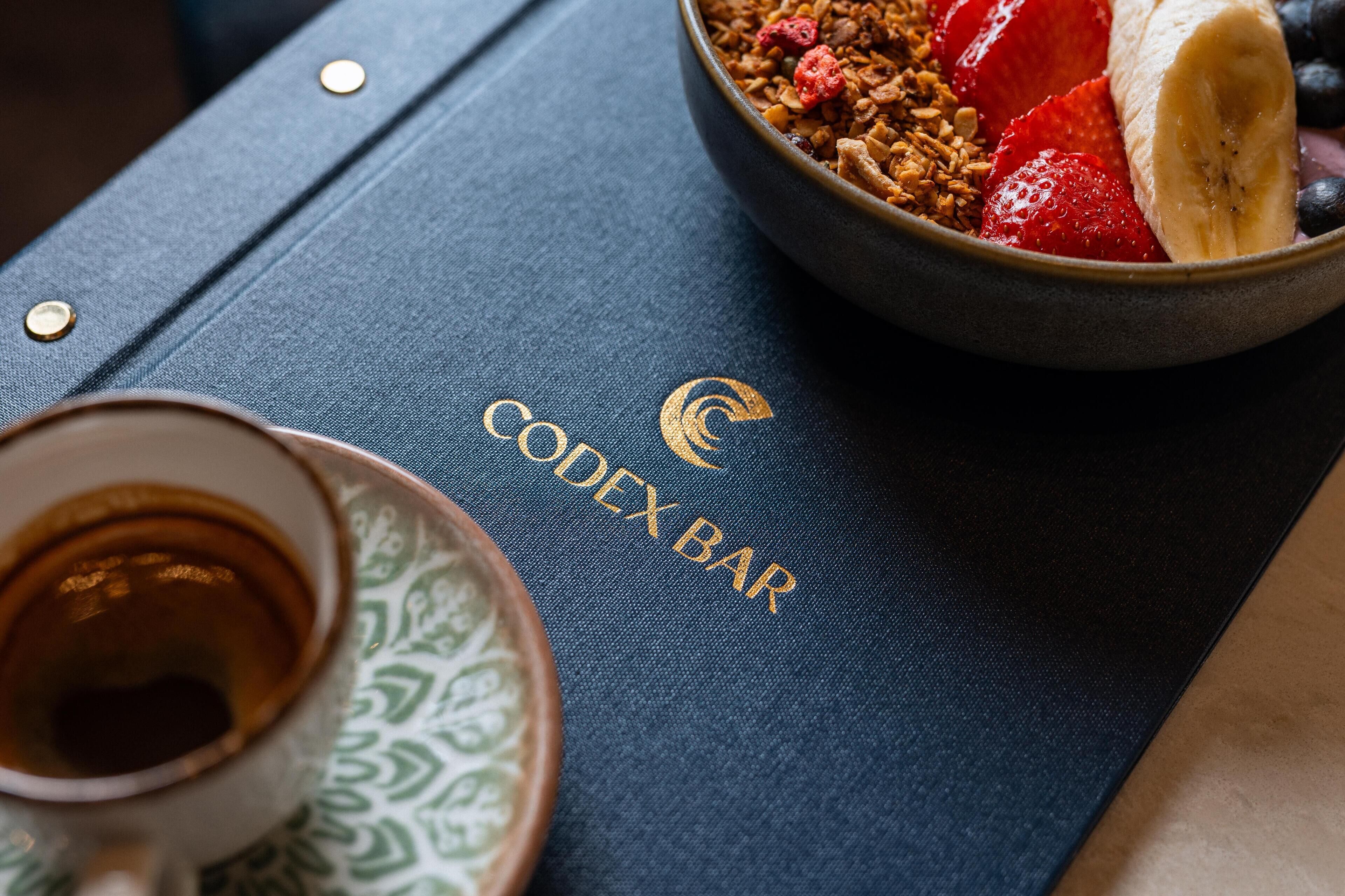 Un libro titulado 'Codex Bar' con un tazón de cereal y fruta encima, una taza de café y un platillo con un diseño verde.