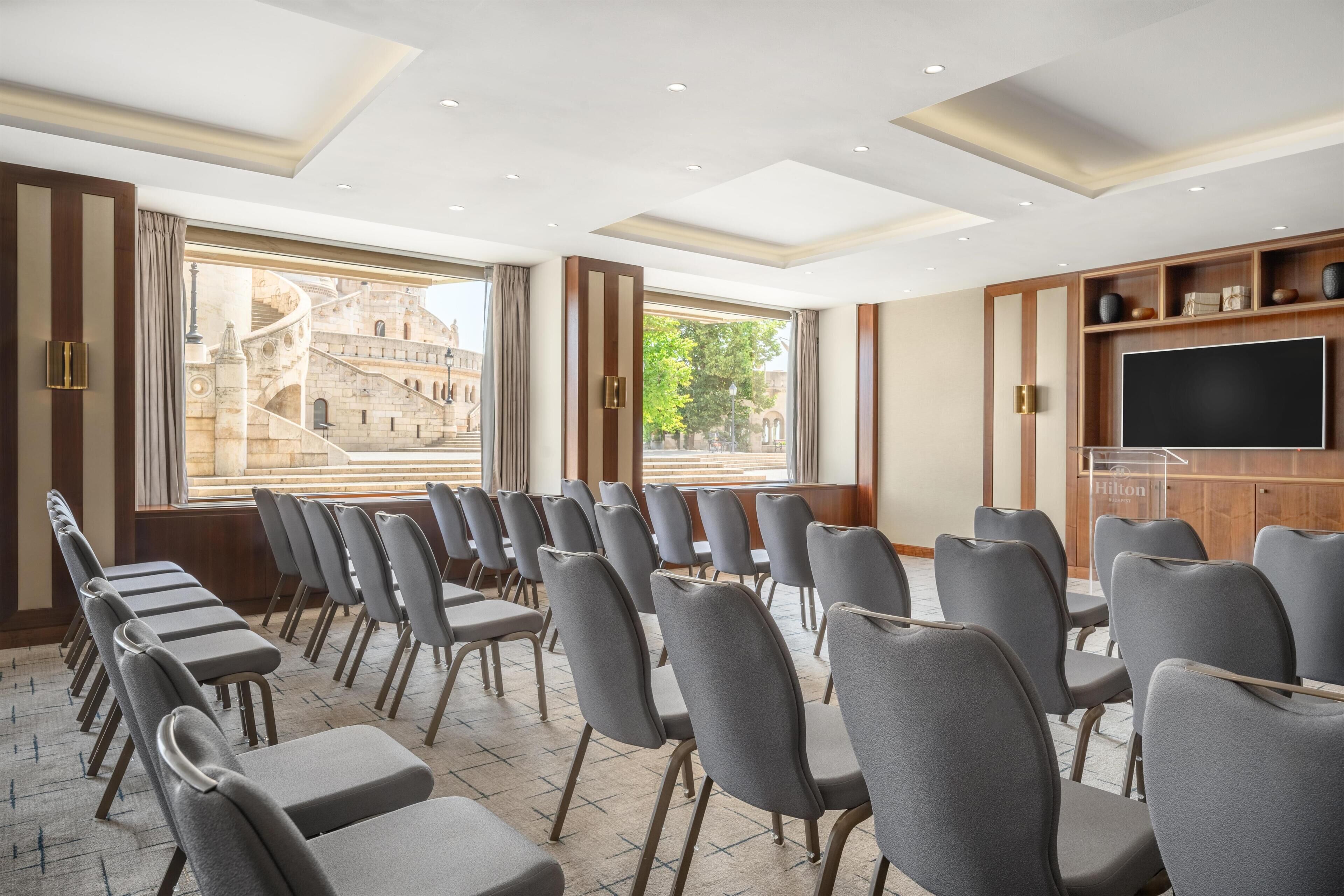 A conference room with rows of chairs and a large screen, large windows with a view of a historic building, and a wooden panel wall with a shelf.