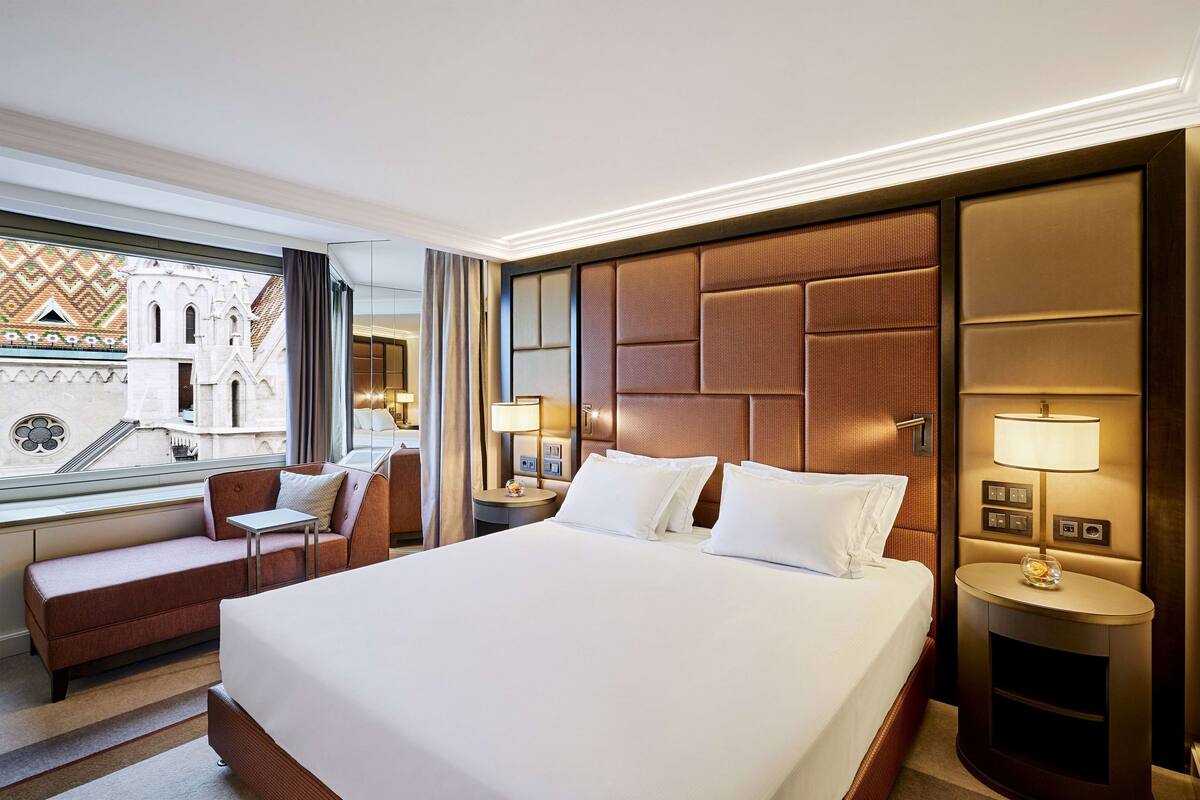 Une chambre d’hôtel avec un grand lit, un canapé, une table de chevet avec une lampe et une vue sur un immeuble à la façade ornée de motifs.