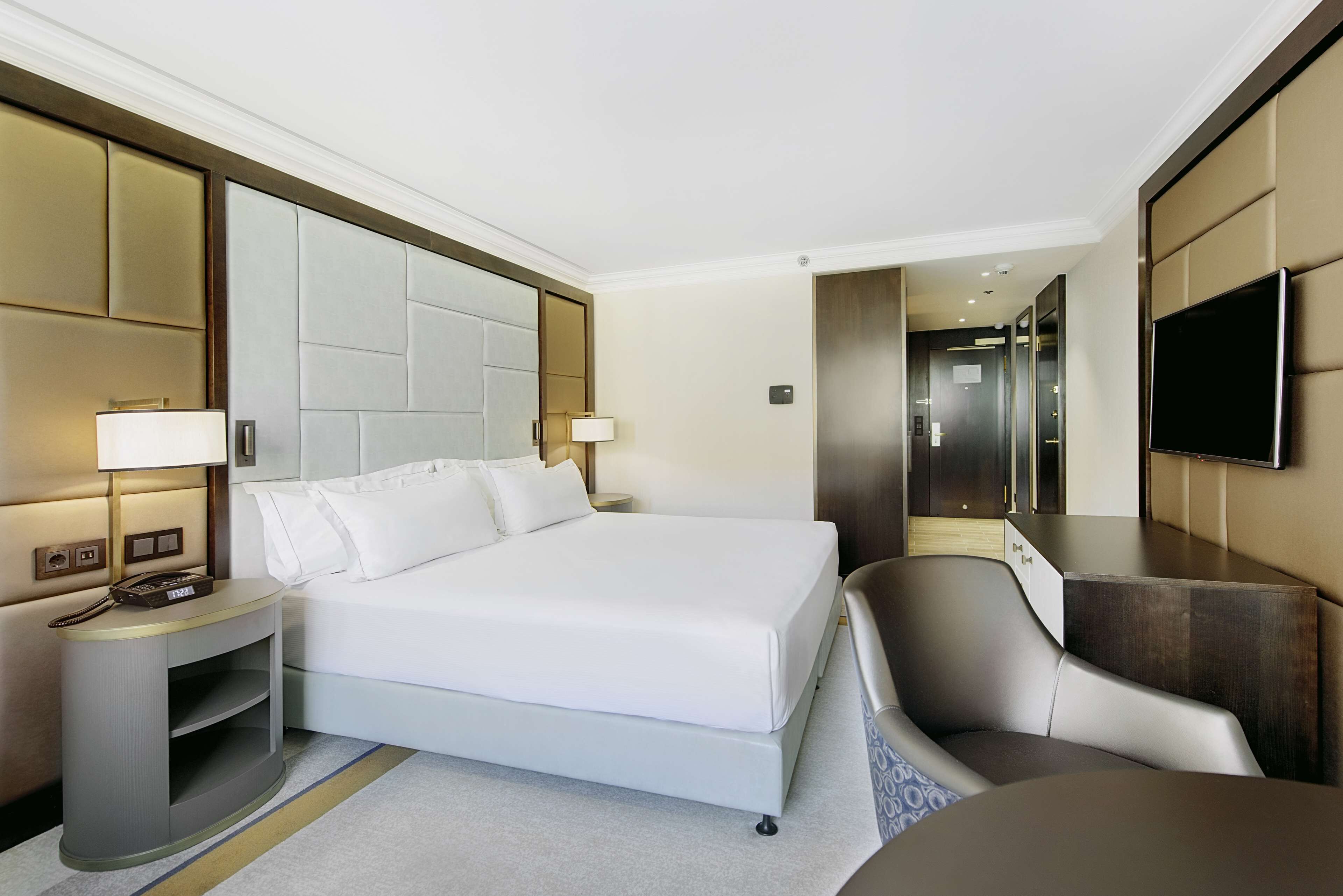 A modern hotel room with a large bed, a desk, a chair, a TV, and a bedside table with a lamp.