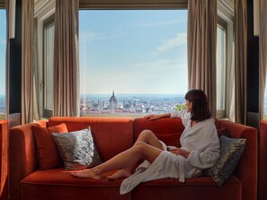 A person sitting on a red sofa with a cityscape view, including a prominent dome structure, through large windows with curtains. - Hilton Budapest (Budapest)