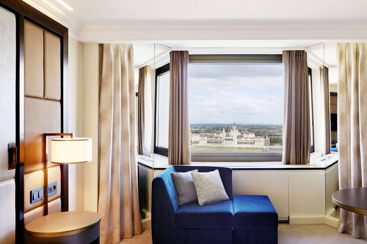 Une chambre d’hôtel avec une grande fenêtre, un canapé bleu, une lampe et une vue sur un bâtiment historique.