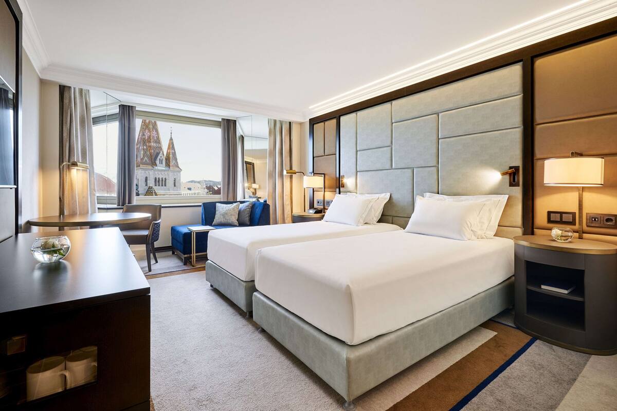 Une chambre d’hôtel avec deux lits, un bureau, une chaise, une petite table et une vue sur un immeuble.
