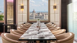 A dining area with a long table set for a meal, leather chairs, and a view of a historic building through a large window. - Hilton Budapest (Budapest)