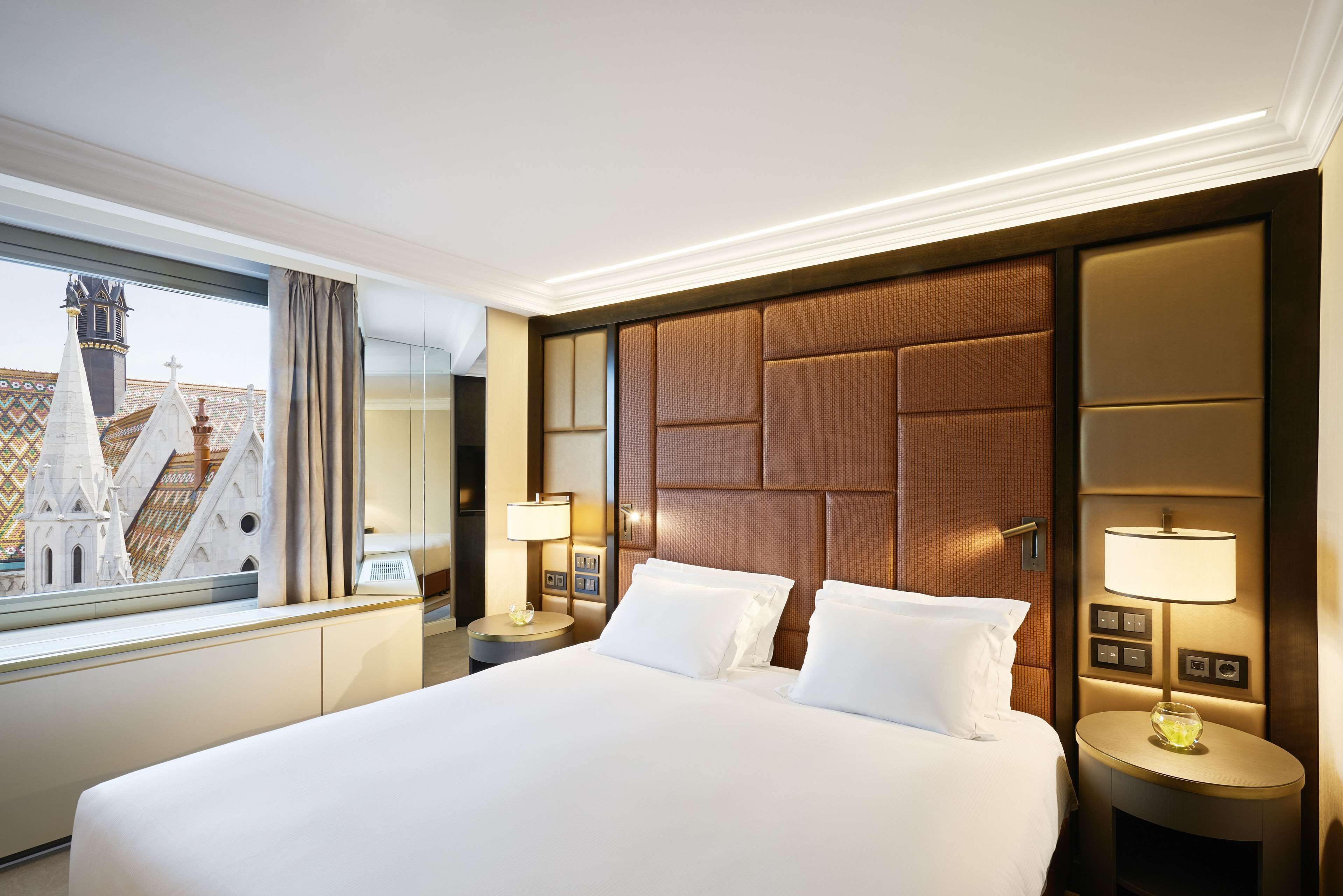 A hotel room with a large bed, a nightstand with a lamp, a window with a view of a building, and a wall with a textured panel.